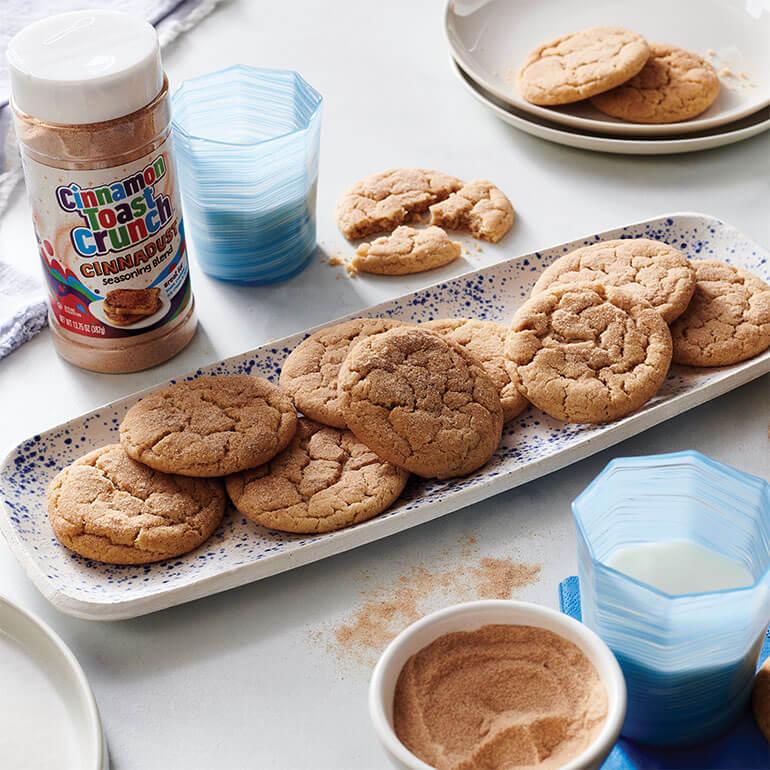 Cinnamon Toast Crunch Cinnadust™ Snickerdoodles on an elongated plate with a jar of Cinnadust™ next to glasses of milk.