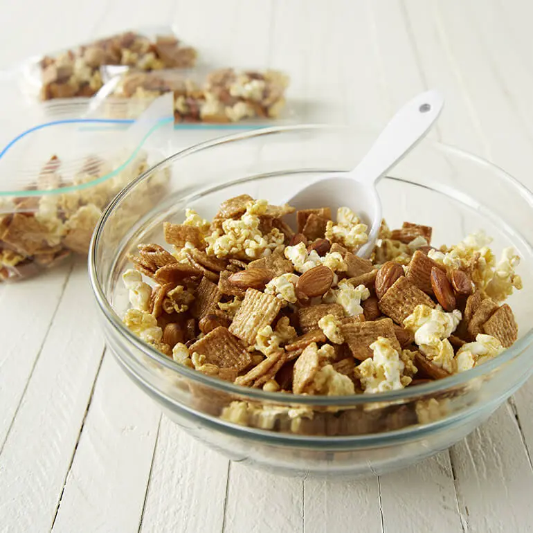 Un bol de Glazed Cinnamon and Popcorn Snack Mix con una cuchara metida en la mezcla y varias bolsas de plástico de la mezcla al fondo.