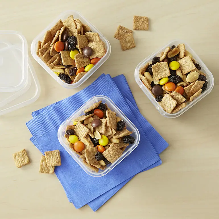 Tres recipientes llenos de Cinnamon Gorp Snack Mix con seis trocitos de cereales derramados en la superficie de abajo y tapas al lado.