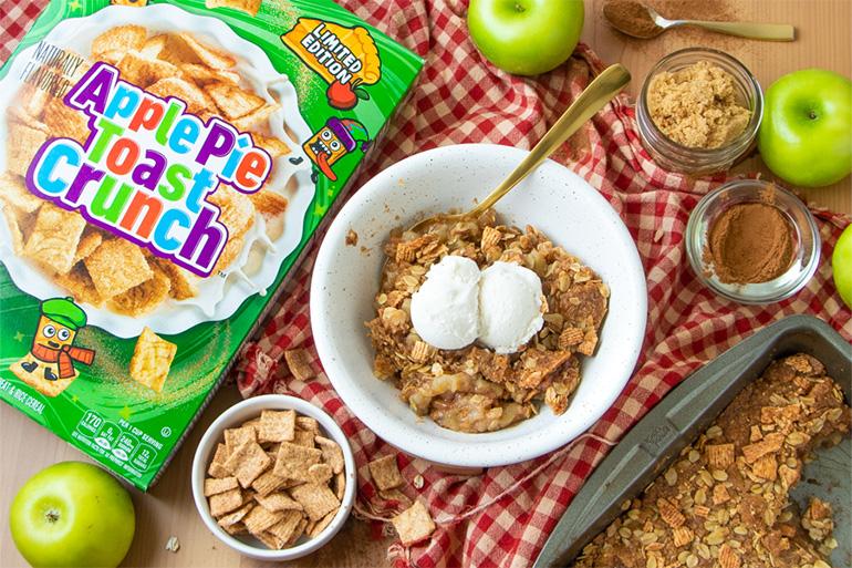 Un bol de Crispy Apple Pie Toast Crunch con una bola de helado encima y una caja de cereales al lado. Los demás ingredientes están eparcidos alrededor.