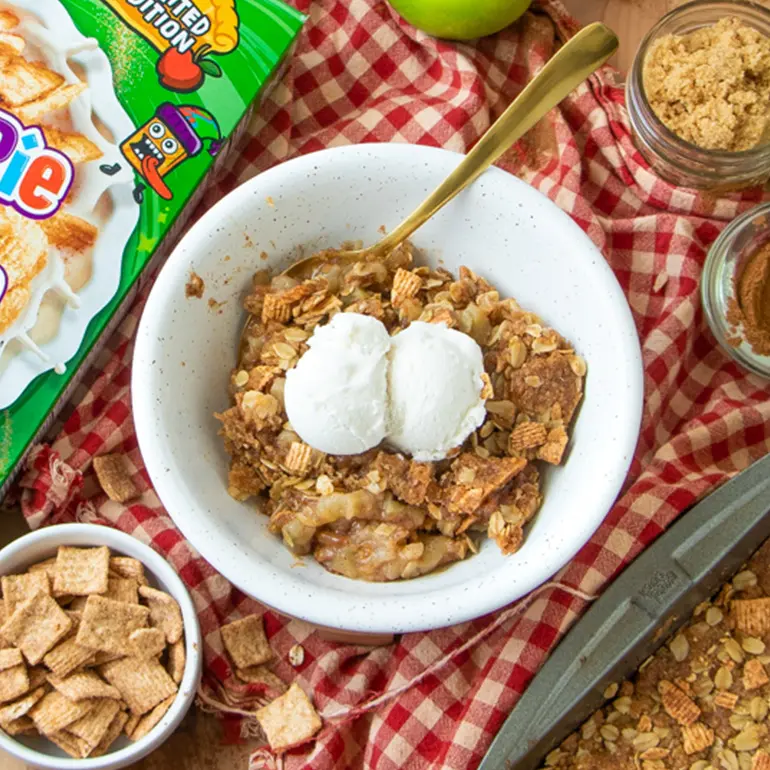 Un bol de Crispy Apple Pie Toast Crunch con una caja de cereales al lado y una bola de helado encima. Hay recipientes de ingredientes alrededor.
