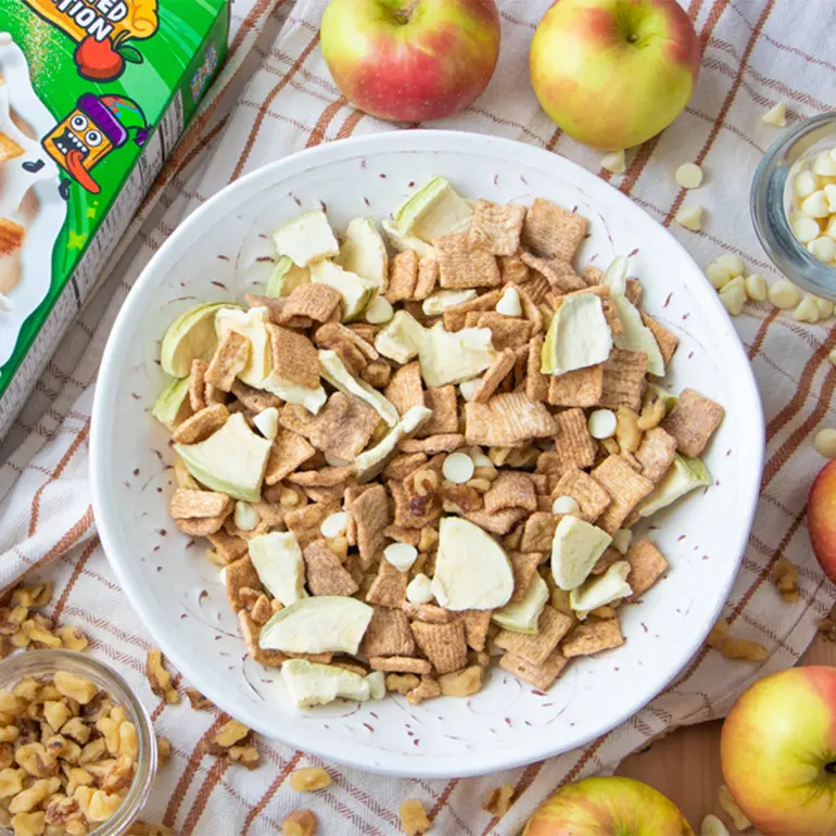 Apple Pie Toast Crunch Snack Mix al lado de una caja del cereal utilizado en la mezcla. Hay cuatro manzanas y otros ingredientes esparcidos alrededor del bol.