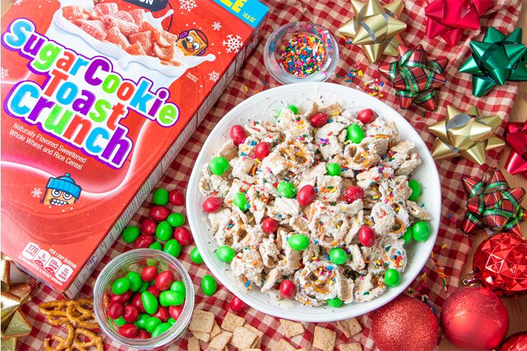Un bol de Sugar Cookie Toast Crunch Snack Mix al lado de una caja de cereales con adornos navideños. Hay tazones con chocolate y chispas de colores.