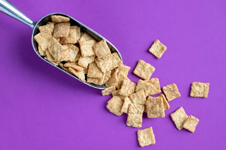 Una pala metálica de mano llena de Cinnamon Toast Crunch. Parte de los cereales se han derramado de la pala y están esparcidos por la superficie.