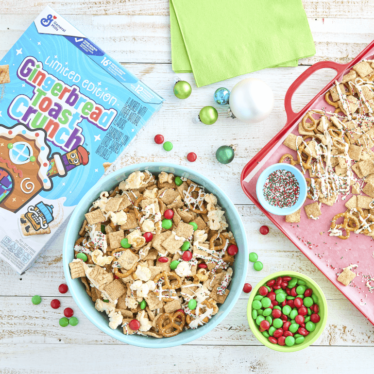 Una caja de Gingerbread Toast Crunch junto a un bol de cereales. Sobre la mesa también hay un pequeño tazón de chocolates confitados y una bandeja de Cereal Snack Mix.