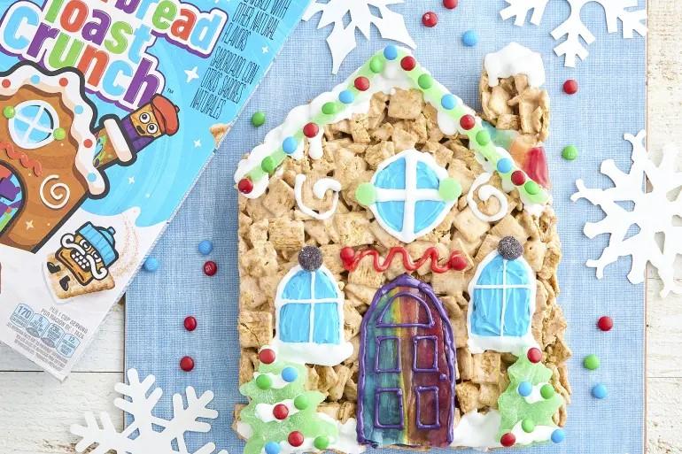 Una caja de Gingerbread Toast Crunch junto a una casa hecha de cereales con puertas, ventanas y nieve hechas de glaseado.