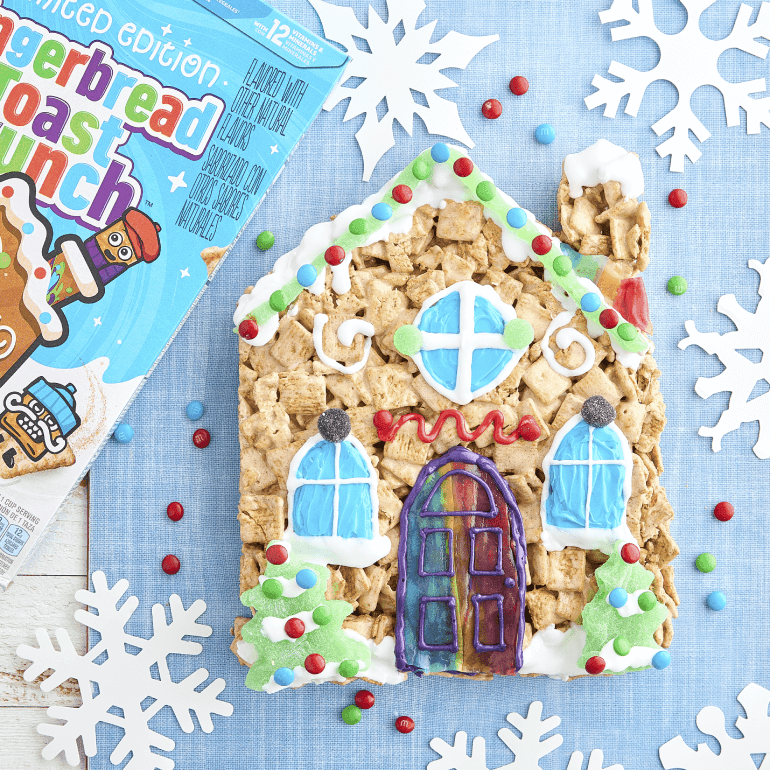 Una caja de Gingerbread Toast Crunch al lado de una casa hecha de glaseado y cereales, con tres ventanas y una puerta.
