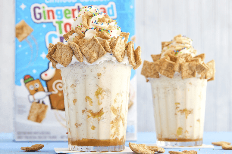 Dos vasos de Gingerbread Toast Crunch Cereal Smoothies decorados elegantemente con una caja de cereales Gingerbread Toast Crunch en el fondo.