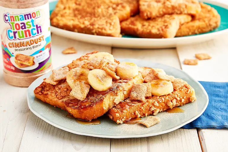 A plate of Cinnamon Toast Crunch™ Cereal-Topped French Toast with a jar of Cinnadust™ behind it.