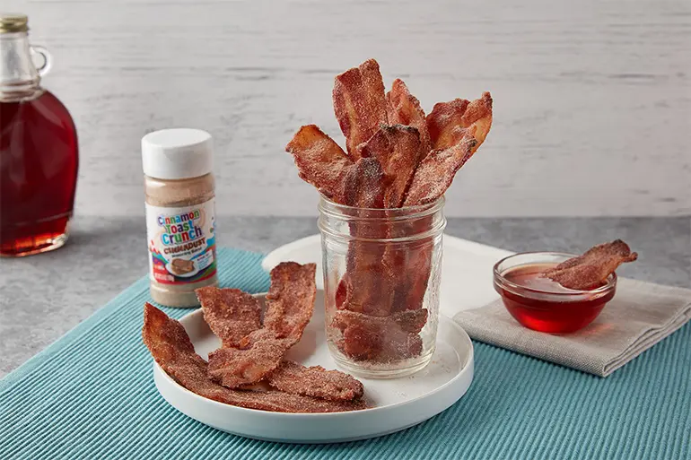 A jar of Cinnadust™ Bacon with some spilled over onto a plate with a jar of Cinnadust™ and syrup behind.