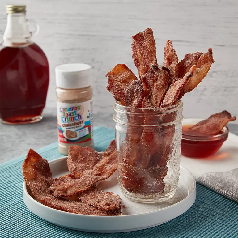 A jar of Cinnadust™ Bacon with some spilled over onto a plate with a jar of Cinnadust™ and syrup behind.
