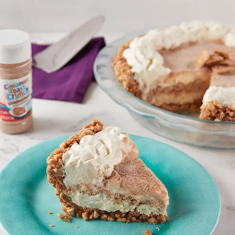 A slice of the Cinnamon Toast Crunch Ice Cream Pie on a plate with the rest of the pie in a dish behind it. A jar of Cinnadust is at the side.