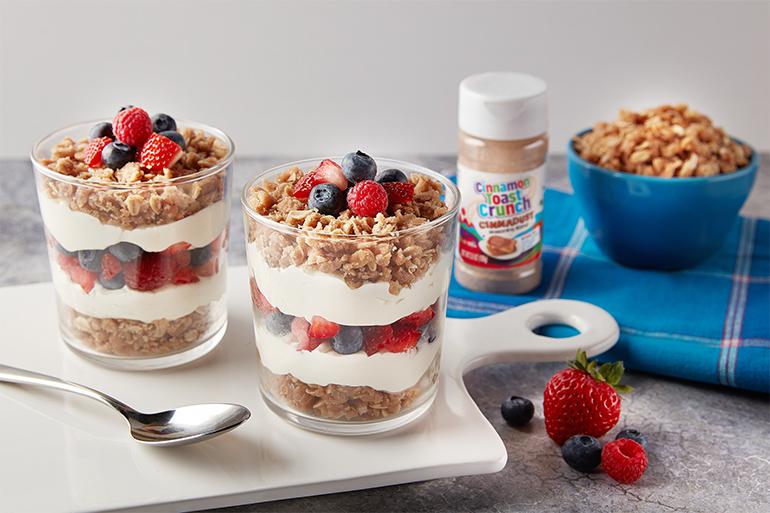 Cinnadust™ Streusel Parfait in 2 clear glass cups with fresh fruit beside them and a jar of Cinnadust™ next to granola.