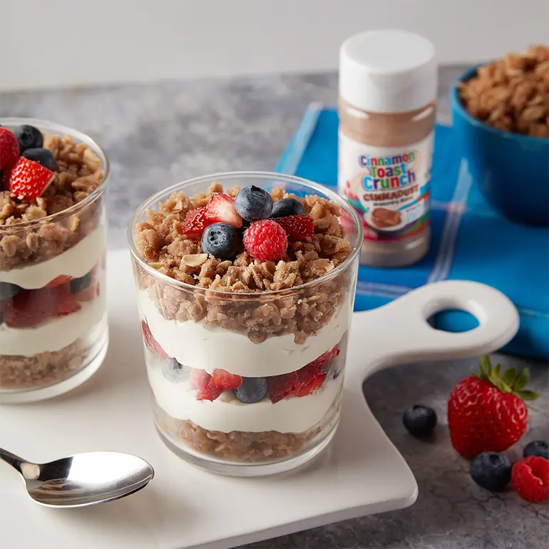 Cinnadust™ Streusel Parfait in 2 clear glass cups with fresh fruit beside them and a jar of Cinnadust™ next to granola.