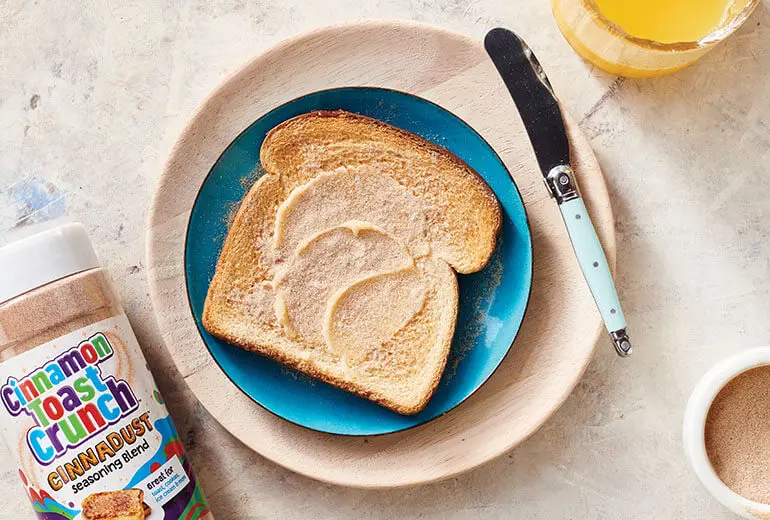 Buttered Toast with Cinnadust™ on a plate with a butter knife, a glass of orange juice and a jar of Cinnadust™ beside it.
