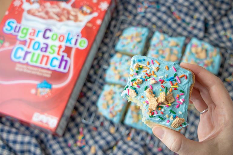 A hand holding a Sugar Cookie Toast Crunch™ Holiday Bar above the Sugar Cookie cereal box.