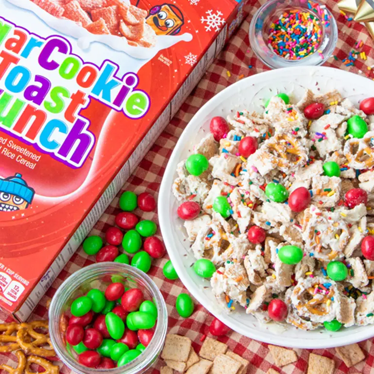 A bowl of the Sugar Cookie Toast Crunch™ Snack Mix surrounded by Christmas decorations and ingredients.